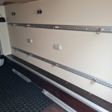 Interior of a cargo trailer showing wall-mounted metal tie-down rails with rings for securing cargo, plus a rubber floor mat and ceiling supports.