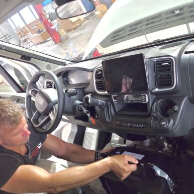 Man in a black shirt sits inside a Fiat van, working on the dashboard near the steering wheel with a large touchscreen display ahead.